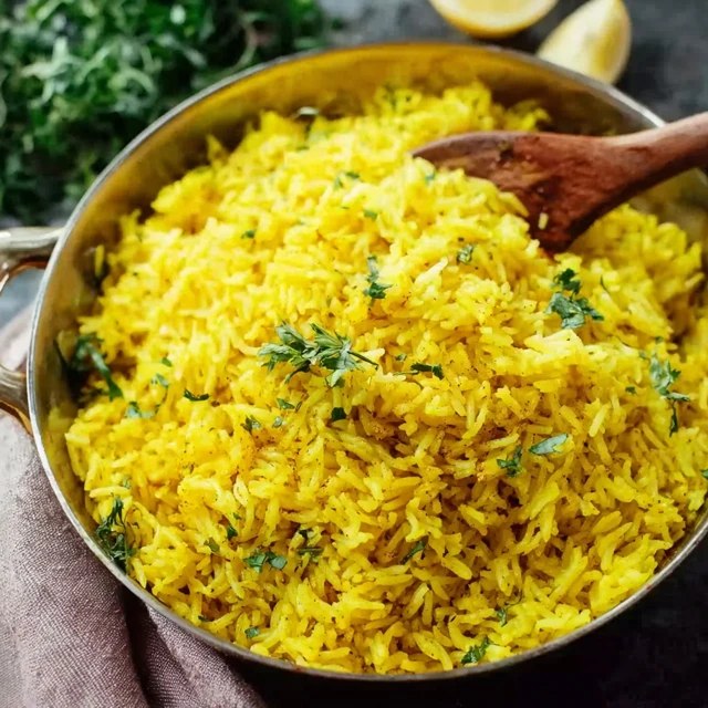 Turmeric rice with herbs in a white serving bowl, vibrant golden-yellow fluffy basmati grains studded with green chopped parsley and cilantro, cumin seed specks, sliced almonds and toasted pine nuts on top, a wedge of lemon on the side