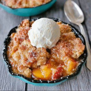 Peach cobbler in a baking dish with golden biscuit topping caramelized brown, juicy bubbling peach filling visible through cracks, large scoop of vanilla ice cream melting on top, cinnamon dust, fresh peach slices garnish